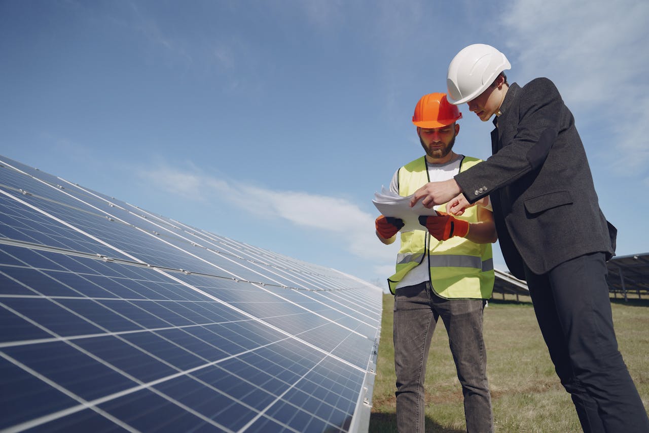 Low angle of young inspector and foreman in hardhats checking documentation against modern solar panels in field